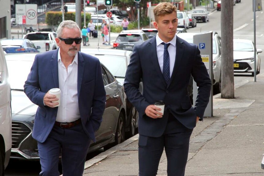 Two men, one older, wearing suits and carrying coffees as they walk up a city street.