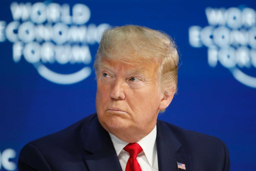 Donald Trump wears a red tie and sits in front of a blue wall with World Economic Forum written on it.