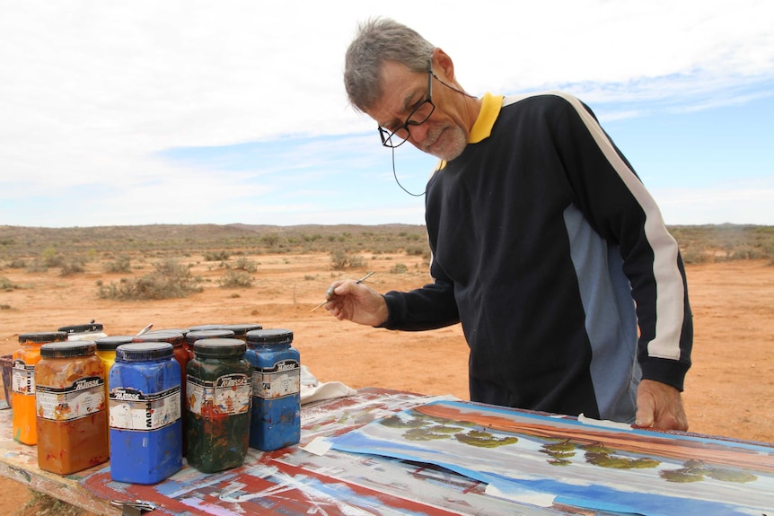 Man holda paintbrush in hand,  looks down at his art work which is a landscape of trees. Tubs of paint sit on the right of table