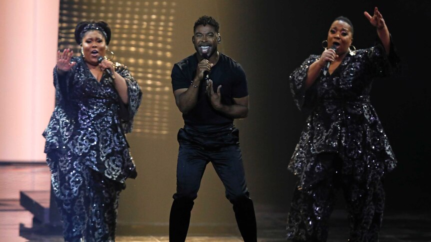 A man performs on stage flanked by two backing singers at Eurovision.