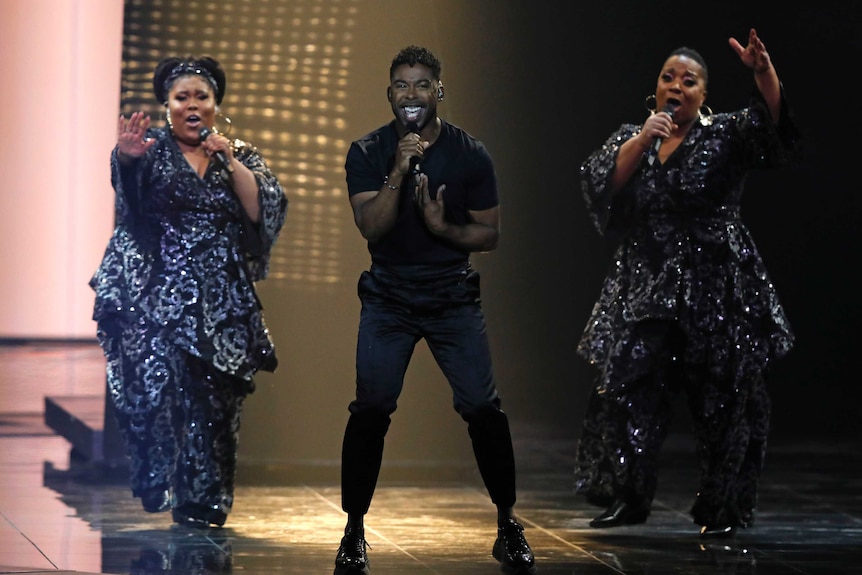 A man performs on stage flanked by two backing singers at Eurovision.