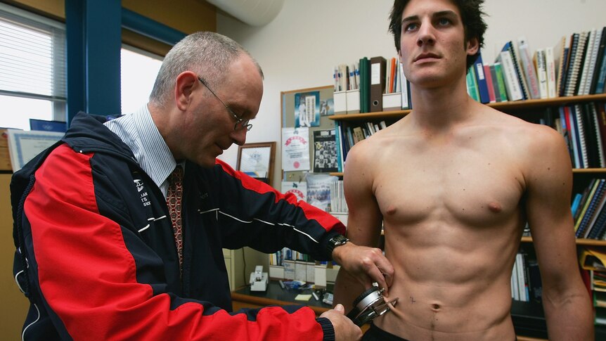An athlete stands straight and looks forward as a doctor uses an instrument to pinch skin on their belly