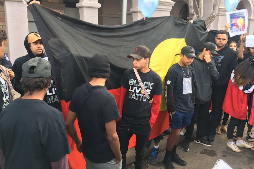 Protestors, some wearing Black Lives Matter t-shirts, stand in front of an Aboriginal flag outside the Kalgoorlie courthouse.