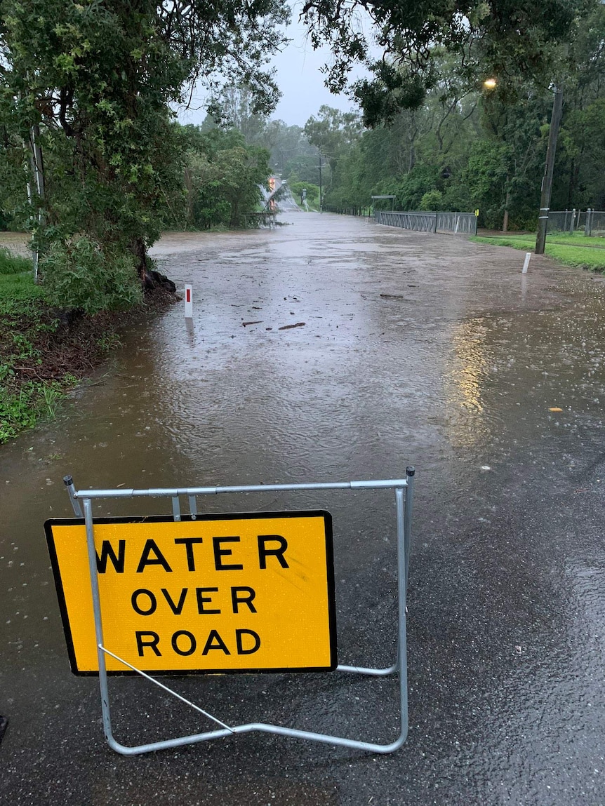 A 'water over road' sign on the road with floodwaters covering the road.