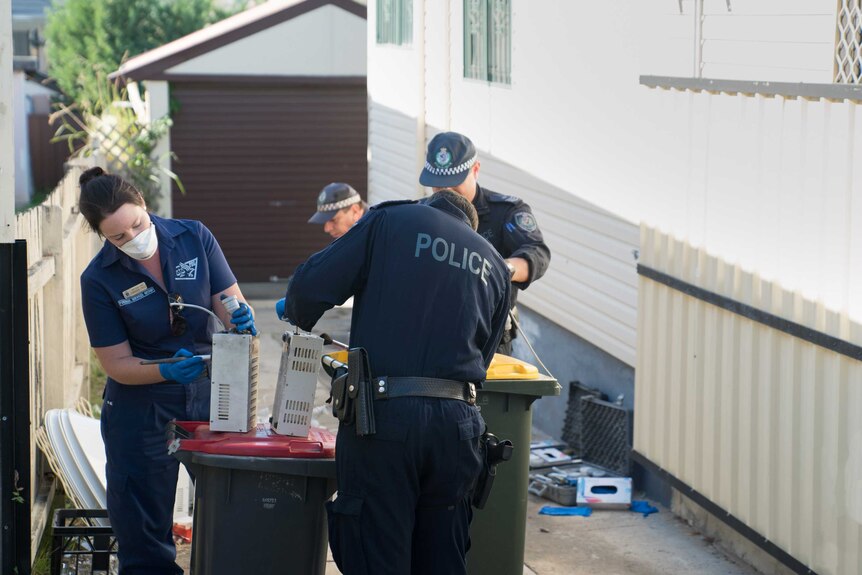 Four police officers wearing blue uniforms dust electrical equipment in a driveway.