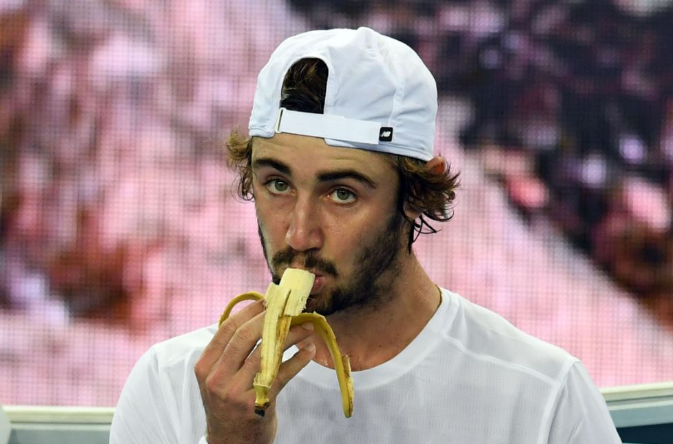 A bearded man wearing a white cap eats a banana.