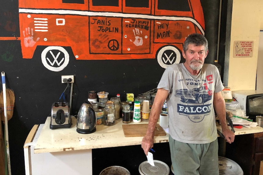 A bearded man in grey t-shirt and shorts stands in front of a toaster, kettle,  kombi van orange painted on wall behind.