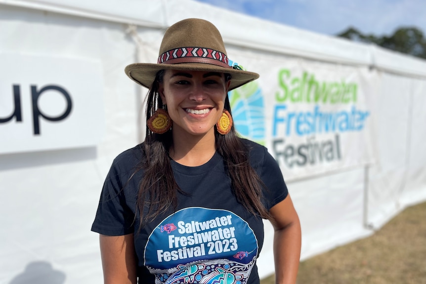 A young Aboriginal woman wearing a t-shirt and wide brimmed hat stands in the sunshine outside a festival tent.