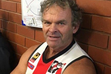 Mullewa Football Club president Mick Wall sits in a changeroom wearing the club's jumper with his arms folded and smiling.