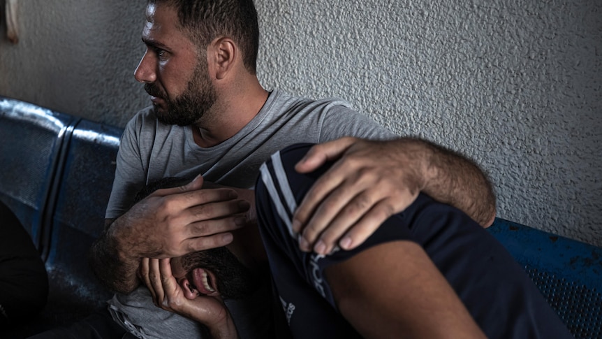 Two men comfort each other as they cry while sitting in a hospital waiting room.