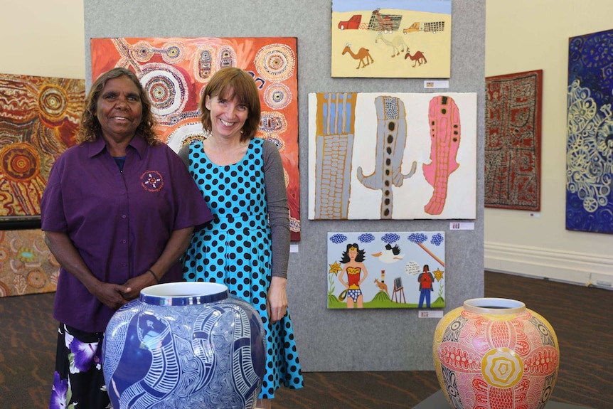 An Aboriginal elder stands alongside a white middle-aged woman surrounded by Aboriginal art in a gallery