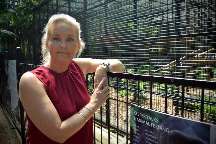 A woman with blonde hair leans over a railing in front of a zoo enclosure.