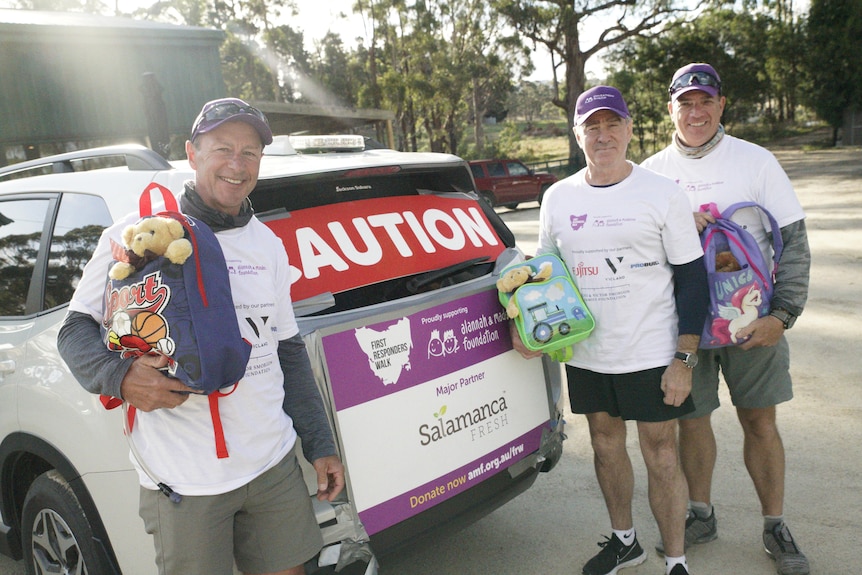 Three men in white t-shirts stand next to a car.