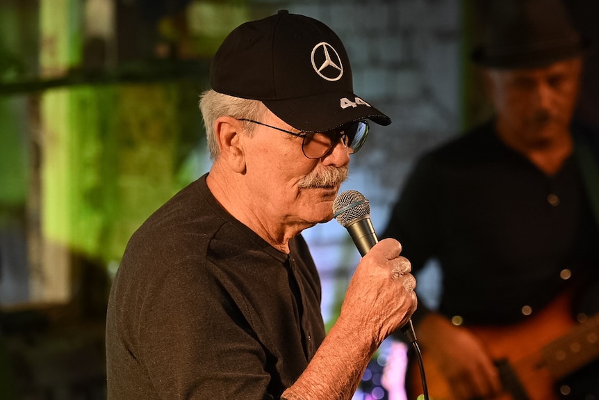 An older man sings into a microphone, wearing a black shirt and black cap with Mercedes logo.