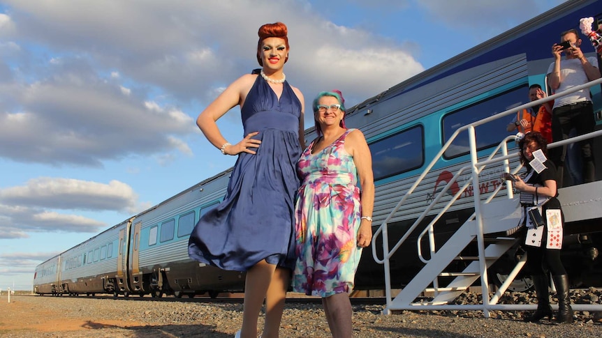 A very tall drag queen stands next to a woman in a colourful dress in front of a train.