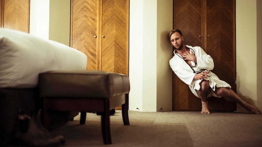 Wearing a dressing gown and holding a glass, Joel Bray slumps against a wall. A double bed is in the foreground.