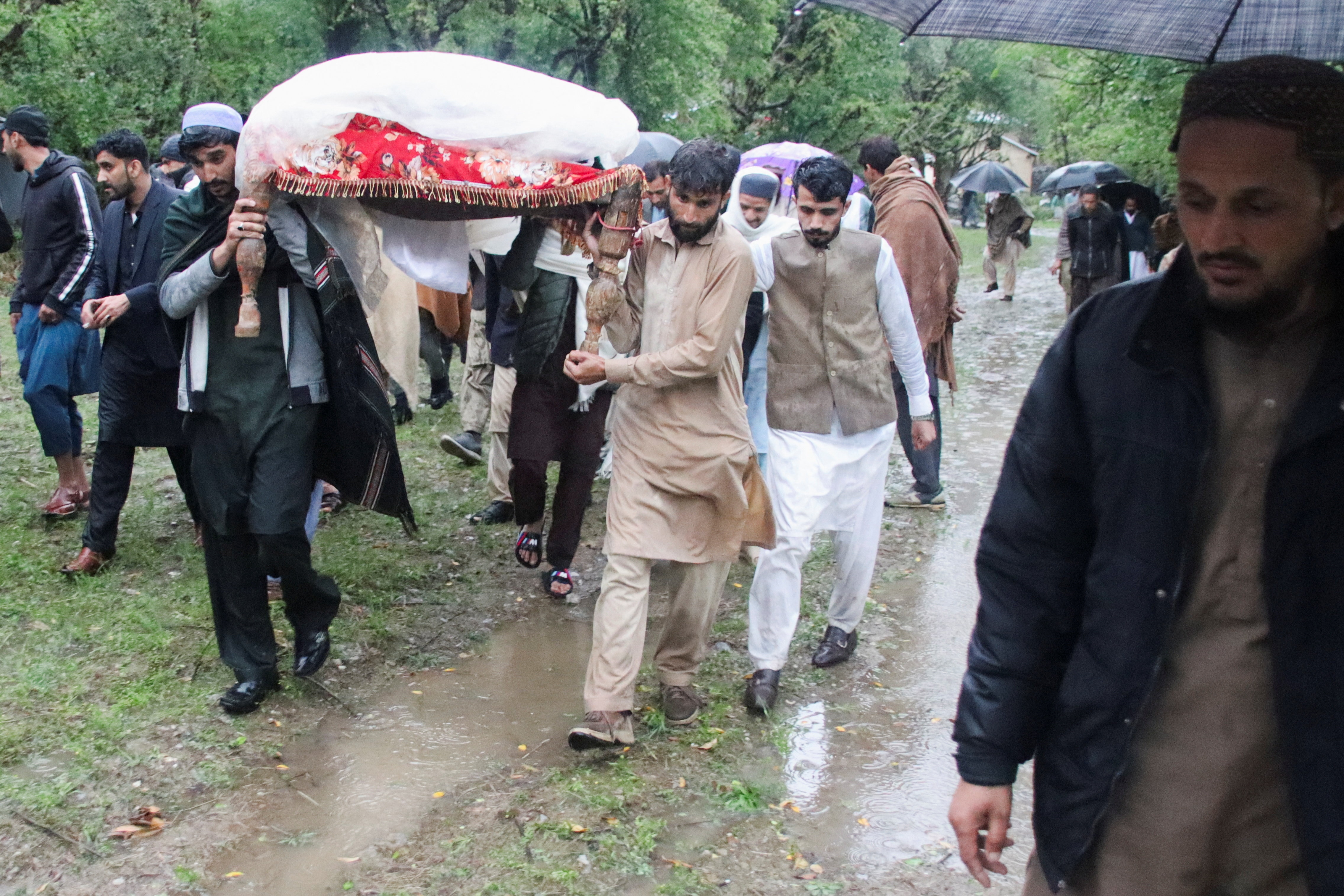 مردان در حالی که آب از پشت سرشان بالا می‌آید، جسد مردی را که با کفن پیچیده شده در جاده‌ای گل‌آلود حمل می‌کنند. 