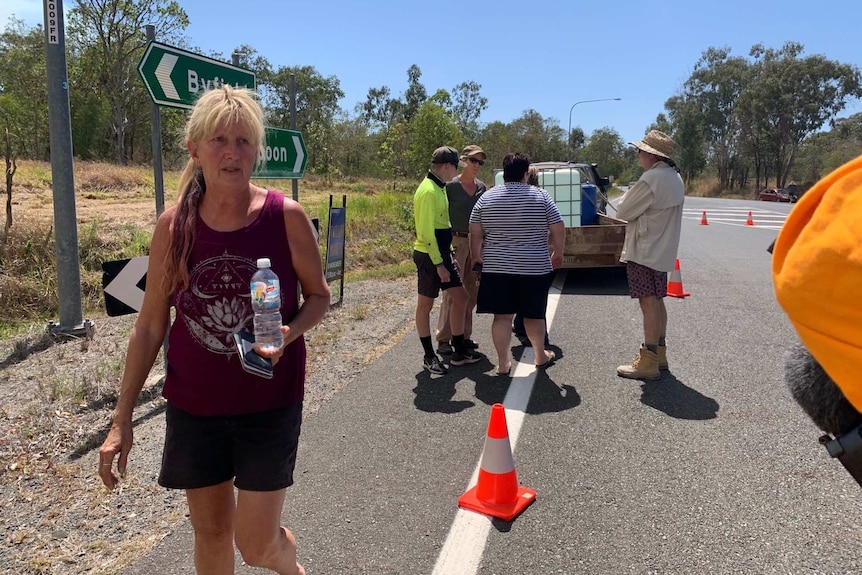 Pauline Allen carries her phone and a water bottle while walking along a road. People are gathered in conversation behind her.