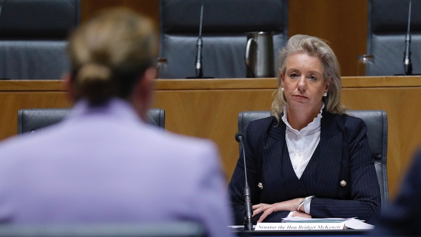 Bridget McKenzie, wearing a dark suit, sits behind a desk and looks at a woman wearing a lilac jacket sitting across from her. 