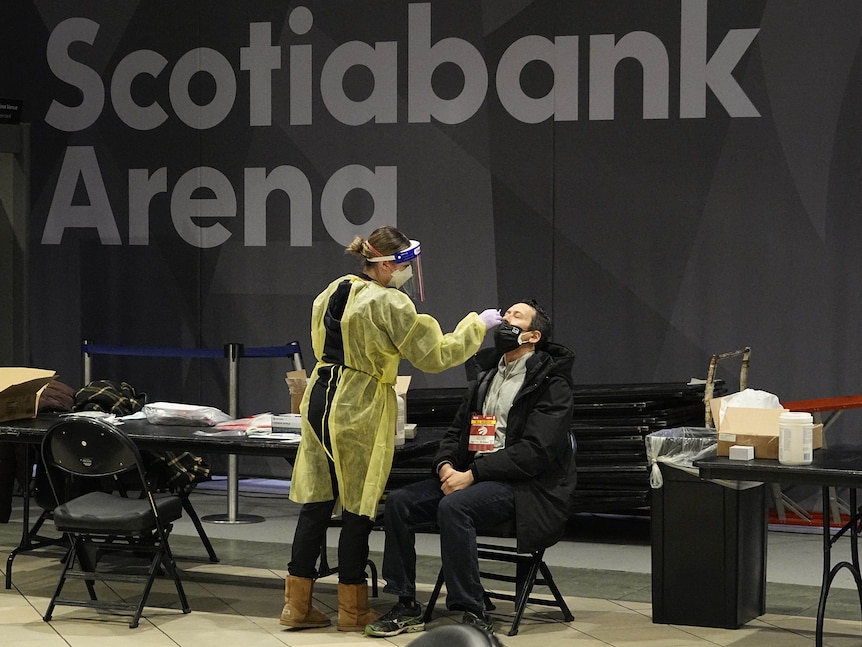 A woman in PPE inserts a sawb into a man's nose as he sits on a chair in front of her, in front of sign saying Scotiabank Arena