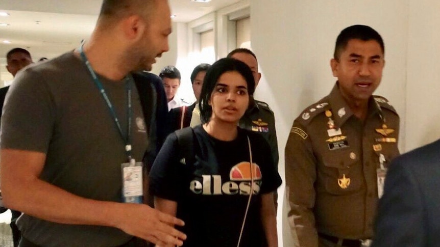 A young woman with a backpack walks down a corridor escorted by officials who are talking to her.