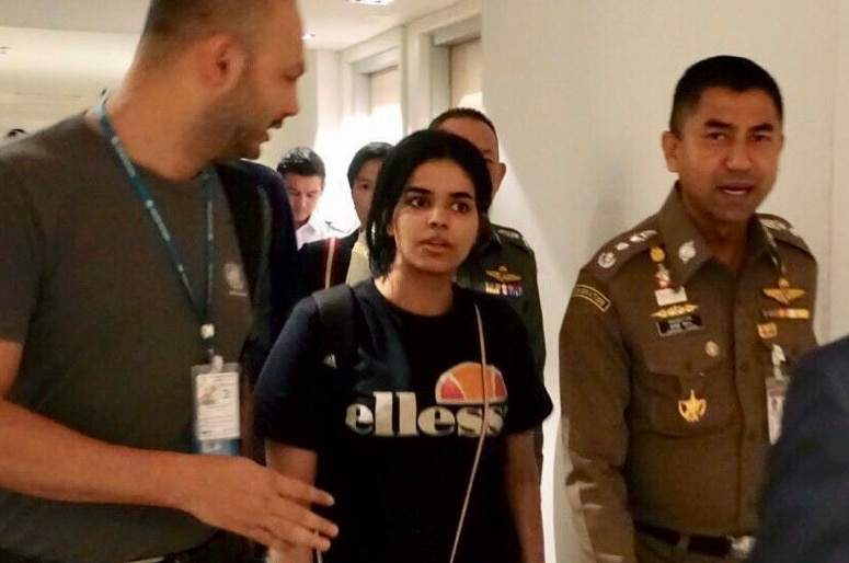 A young woman with a backpack walks down a corridor escorted by officials who are talking to her.