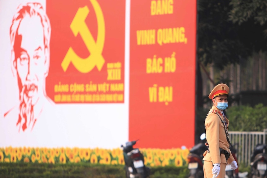 A Vietnamese guard in uniform wears a mask and stands in front of a poster of Ho Chi Minh.