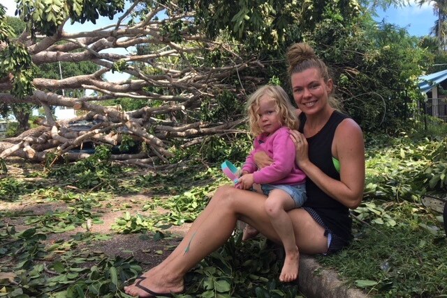 A mum and her daughter sit on the curb, with a massive fallen tree behind them.