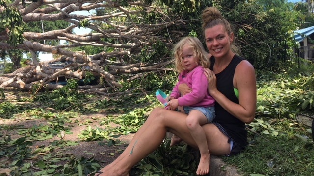 A mum and her daughter sit on the curb, with a massive fallen tree behind them.