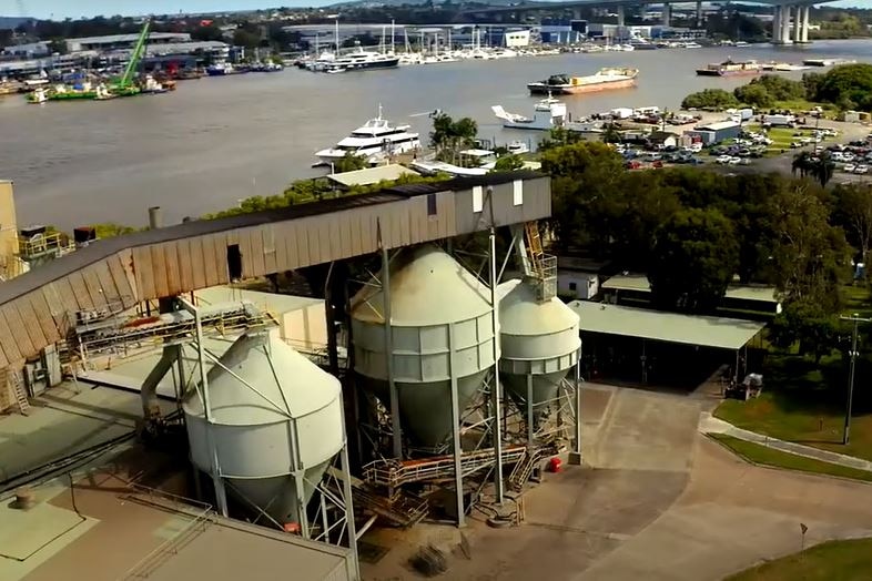 An industrial facility on the Brisbane river, pictured from a height. In the foreground are three silo-type structures.