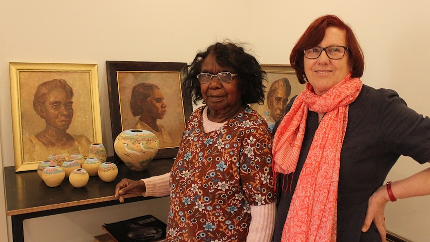 The pair are pictured indoors, standing next to a shelf with portraits displayed