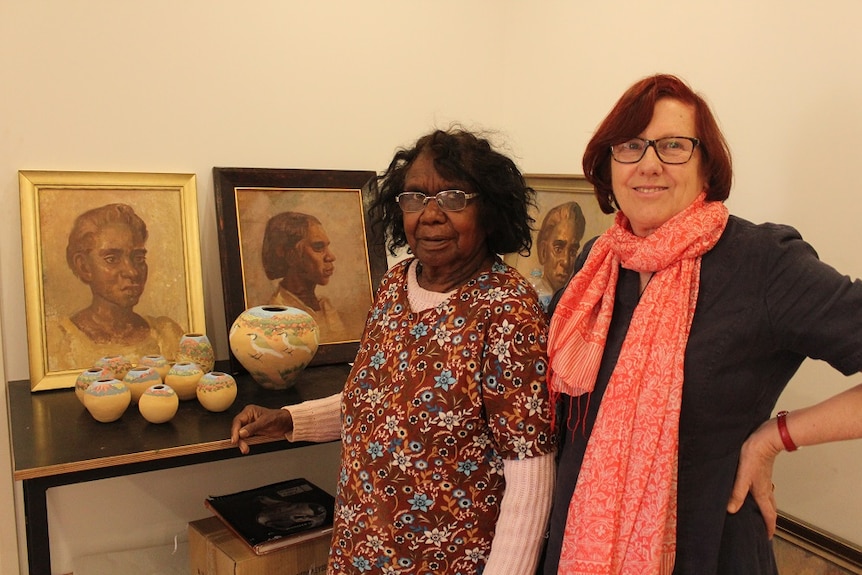 The pair are pictured indoors, standing next to a shelf with portraits displayed