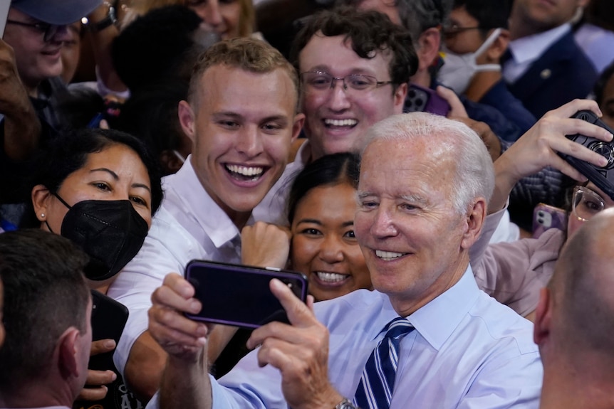 Holding a phone Joe Biden takes a selfie as a crowd of people behind him smile. 