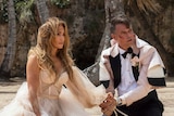 A Latina woman with long honey hair in a wedding dress sits exasperatedly on a beach with bridegroom in white and black tux.