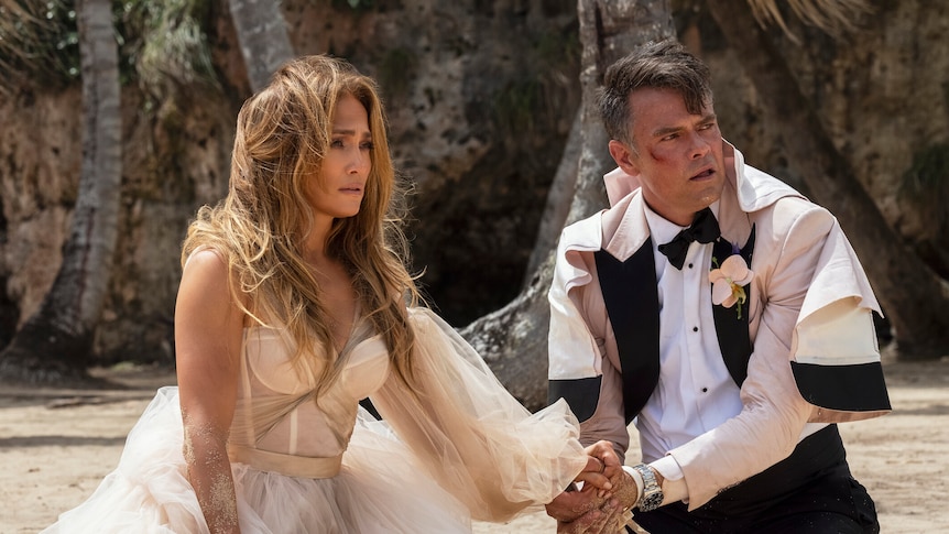 A Latina woman with long honey hair in a wedding dress sits exasperatedly on a beach with bridegroom in white and black tux.