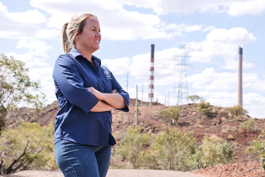 A woman in a blue work shirt standing outside with smoke stacks visible in the background.
