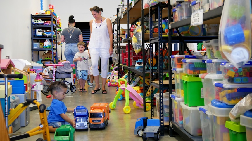 A mother and son walk down the aisle of a toy library while a young boy plays with toy trucks