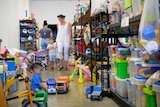 A mother and son walk down the aisle of a toy library while a young boy plays with toy trucks