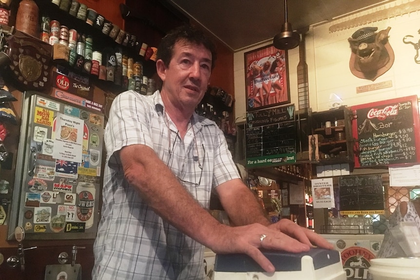 A publican stands behind the bar of a typical country pub with walls and fridges adorned with paraphernalia