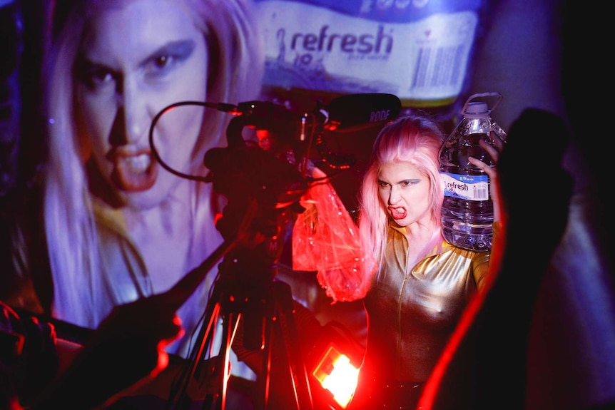 Woman dressed like 80s female wrestler, with pink hair and blue eyeshadow, grimaces to camera - her face projected behind.