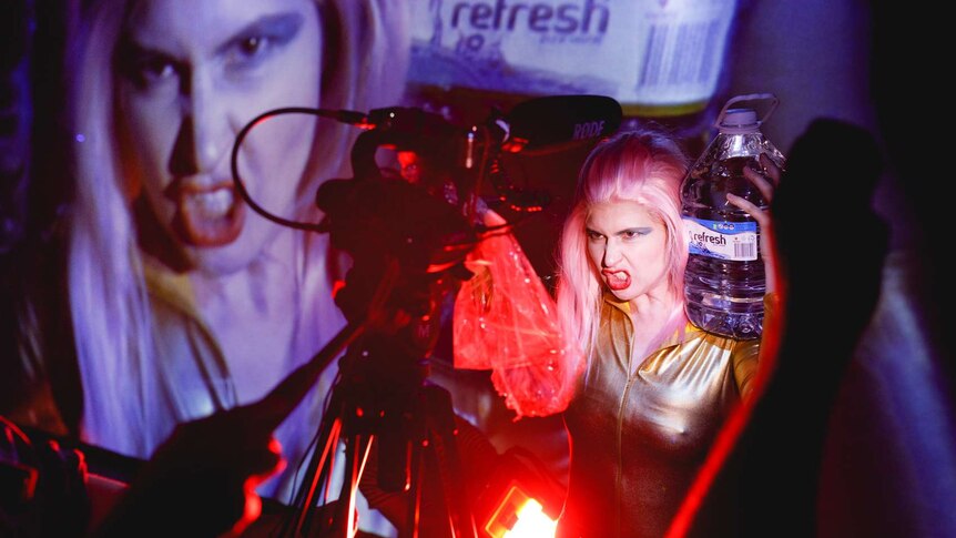 Woman dressed like 80s female wrestler, with pink hair and blue eyeshadow, grimaces to camera - her face projected behind.