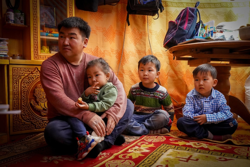 A man in red long-sleeved Tshirt sits with a child on his lap and two others sitting cross legged next to him