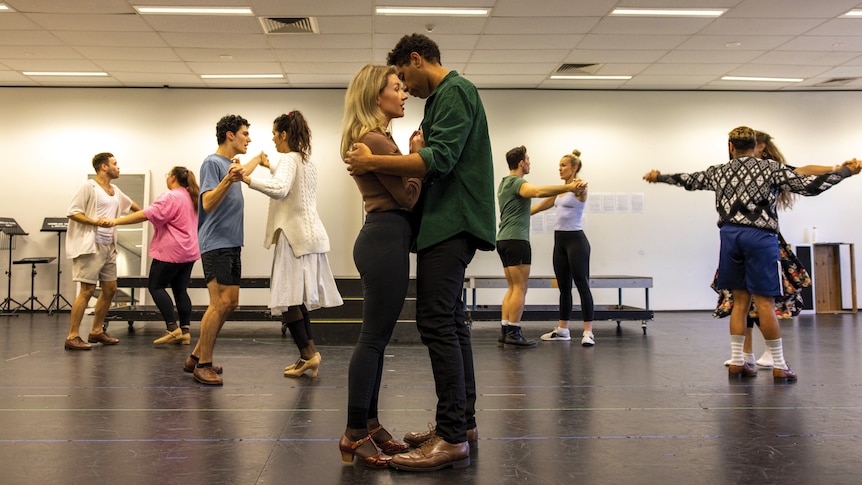 White woman and Indigenous Australian man stand close together in a rehearsal room while four couples dance around them