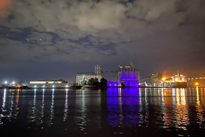 Silos are lit up with purple lights on a body of water with clouds in the sky at night.