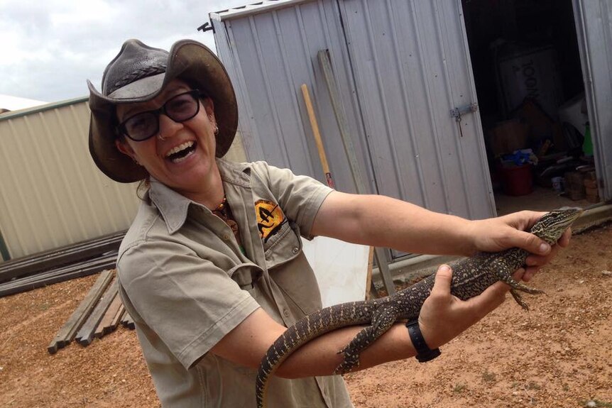 Michelle Jones wears khaki clothes, a leather bush hat and holds a large lizard. She's got a big smile on her face.