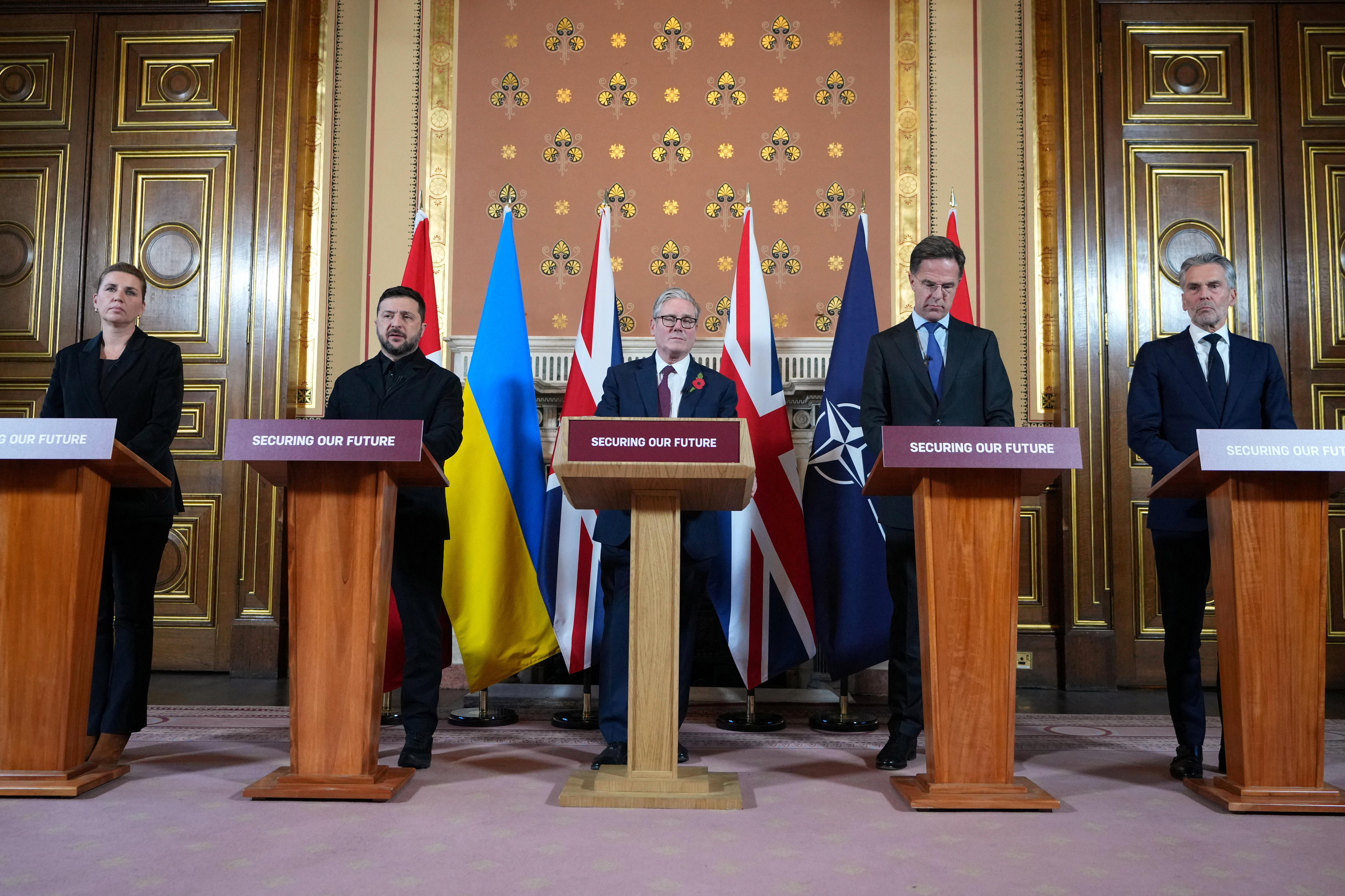 A group of people stand at a row of lecturns in front of brightly coloured flags in a grand room.
