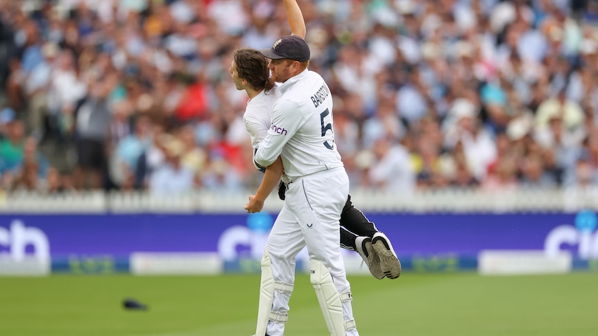A cricket player in whites carries a protester off the ground.