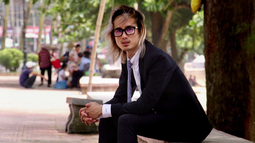 Jack Ta, wearing glasses with died blond and black hair, sits in a street in Vietnam wearing a suit.