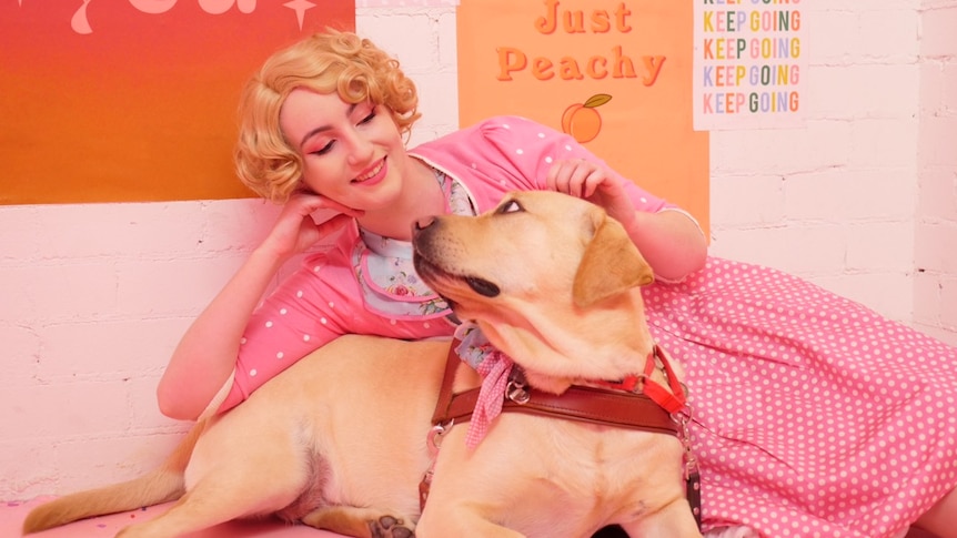 A woman wears a pink robe and lays next to her guide dog. She is smiling