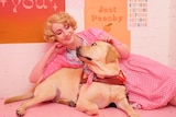 A woman wears a pink robe and lays next to her guide dog. She is smiling